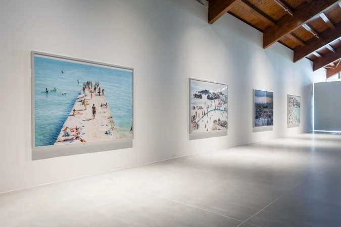 Art gallery exhibition featuring framed beach scene photographs with people, pier, and blue ocean, complemented by the gallery's modern architecture.
