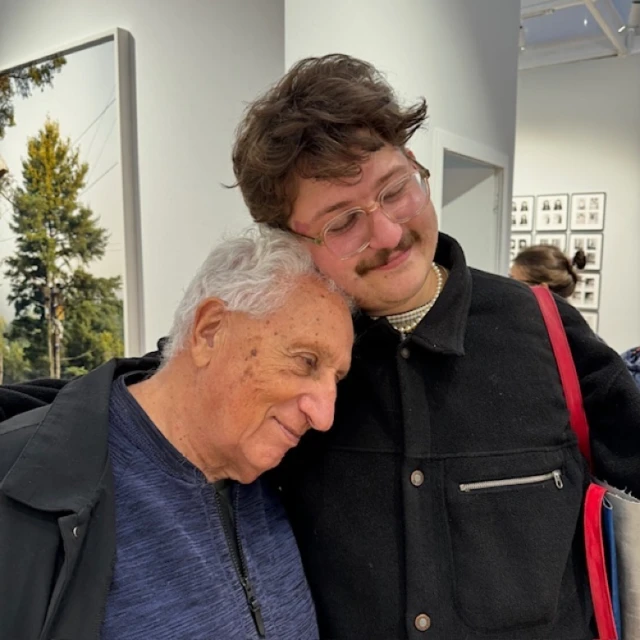 An elderly man with white hair is embracing a younger man with glasses and a mustache inside an art gallery with artwork on the walls.