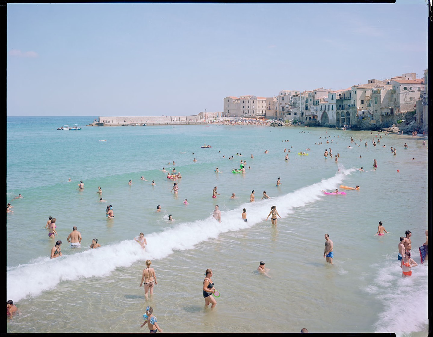 Crowd of people swimming and wading in the sea at a beach, with a small wave rolling in and buildings on the coast in the background.