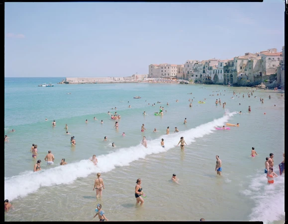 Crowd of people swimming and wading in the sea at a beach, with a small wave rolling in and buildings on the coast in the background.