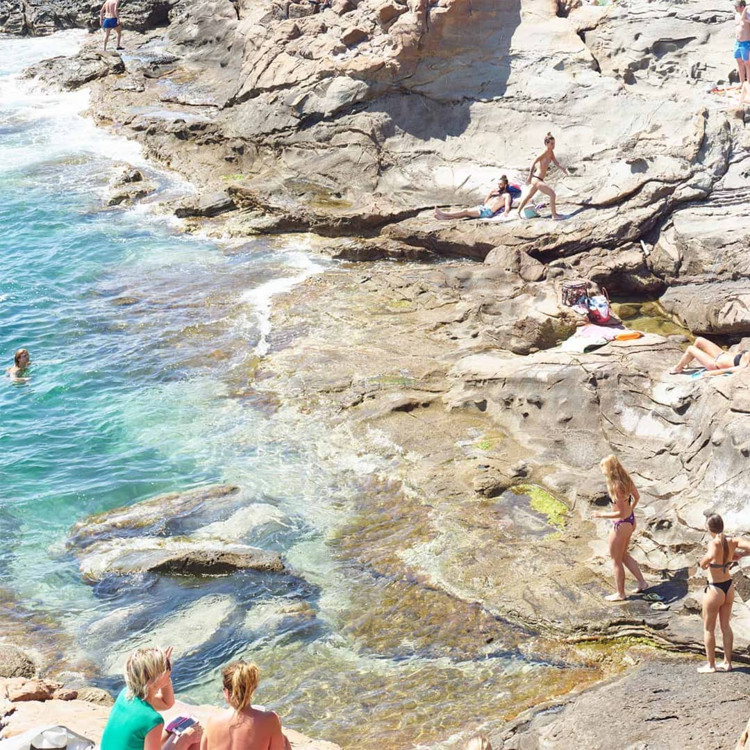 People enjoying the rocky beach and clear turquoise water on a sunny day. Adults are sunbathing, swimming, walking, and relaxing by the ocean.
