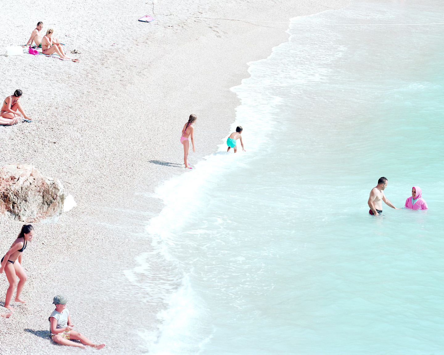Aerial view of people on a pebble beach and in turquoise water, with adults and children in swimwear enjoying a sunny day.