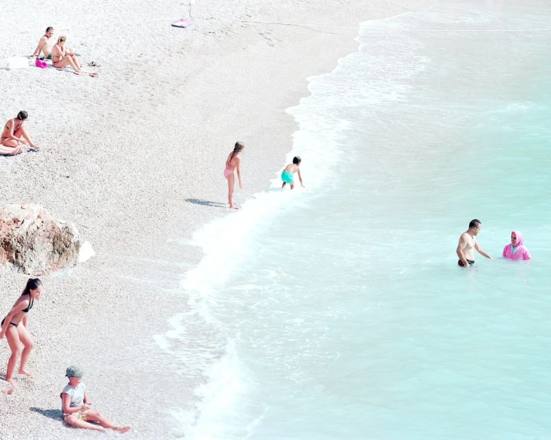 Aerial view of people on a pebble beach and in turquoise water, with adults and children in swimwear enjoying a sunny day.