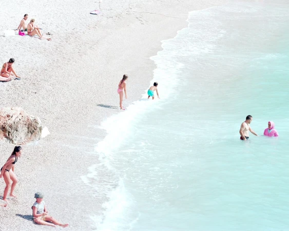 Aerial view of people on a pebble beach and in turquoise water, with adults and children in swimwear enjoying a sunny day.