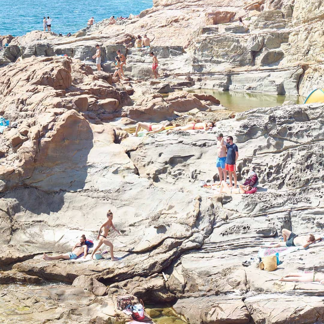 Sunbathers relax on the rocky coast with tide pools. People lounge on the rock formations, some swim in the ocean, enjoying the sunny weather.