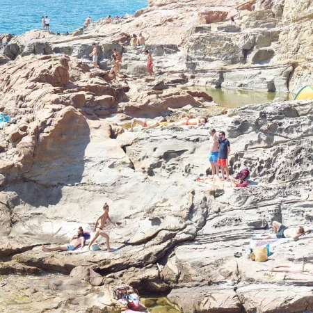 Sunbathers relax on the rocky coast with tide pools. People lounge on the rock formations, some swim in the ocean, enjoying the sunny weather.