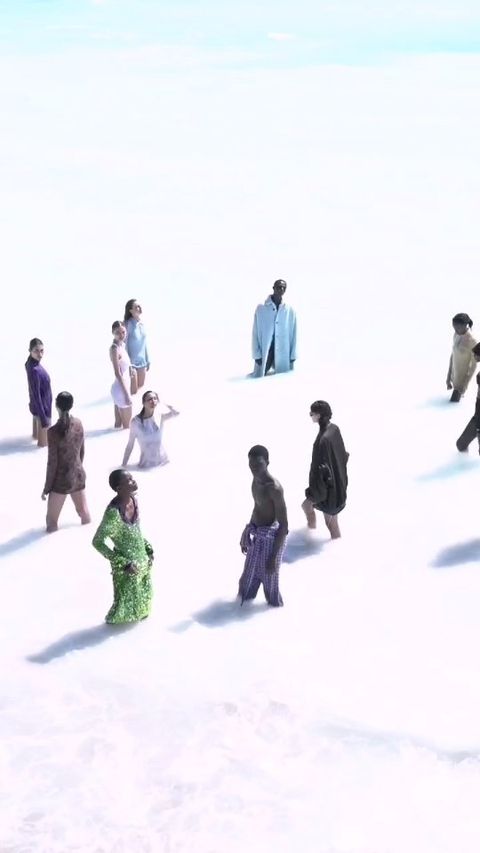 Group of diverse men and women stand in white field under light blue sky, wearing assorted casual clothing in an outdoor fashion editorial.