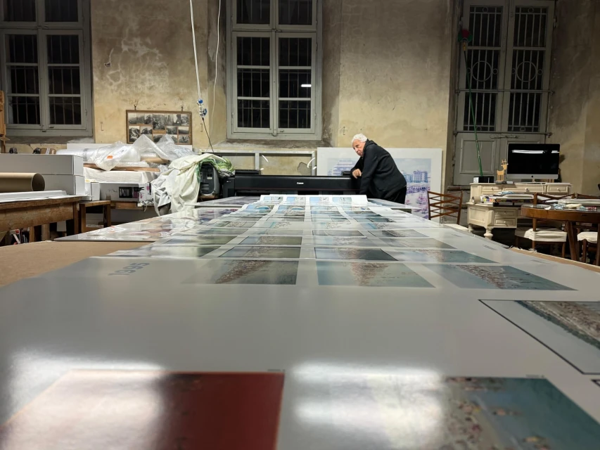 Adult is positioned at a large format printer amid prints in a cluttered photo studio with large windows and low lighting.