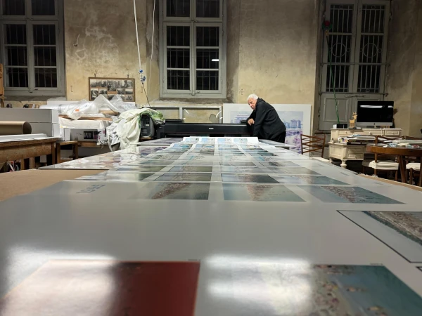 Adult is positioned at a large format printer amid prints in a cluttered photo studio with large windows and low lighting.