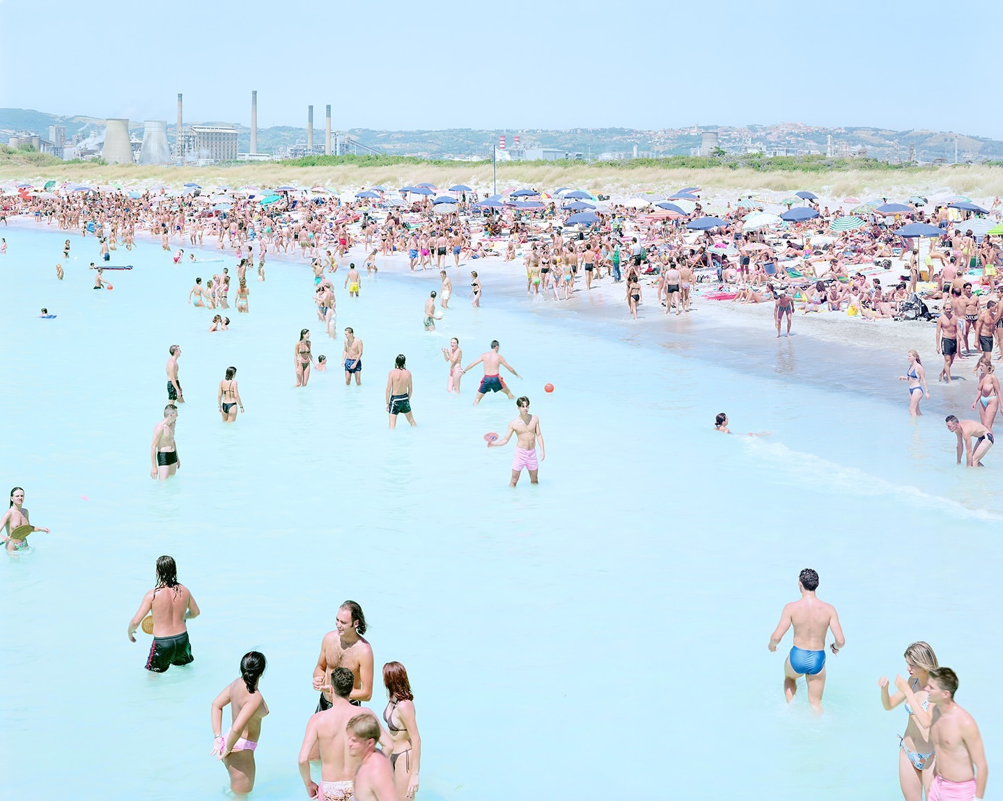 A large crowd of people enjoys a sunny day at the beach, with many swimming and sunbathing in the turquoise water.