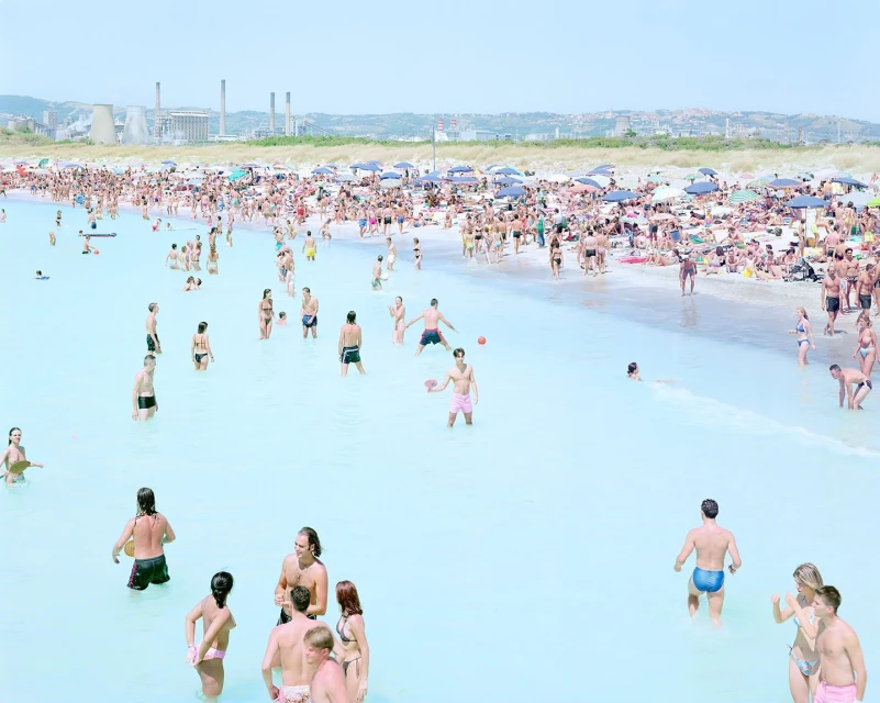 A large crowd of people enjoys a sunny day at the beach, with many swimming and sunbathing in the turquoise water.