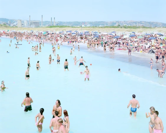 A large crowd of people enjoys a sunny day at the beach, with many swimming and sunbathing in the turquoise water.