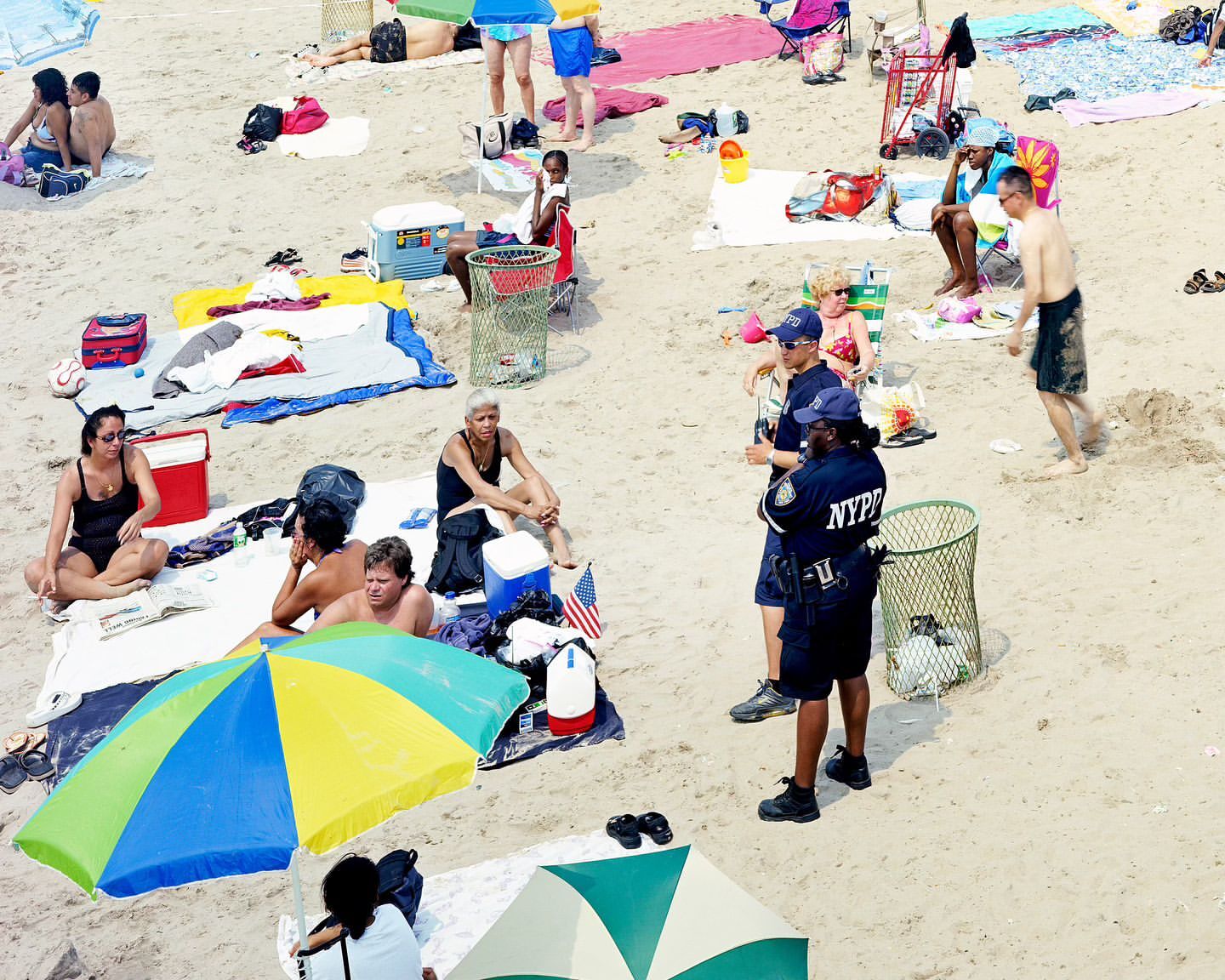 Crowded beach scene with sunbathers, umbrellas, and two NYPD officers, capturing a busy summer day from an elevated perspective.
