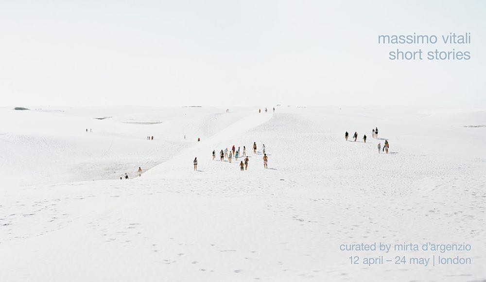 A group of people stroll across light sand dunes under a bright sky, captured in a wide shot showcasing minimalist aesthetic.