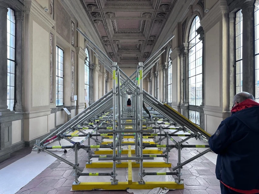 Interior view of a long hall with scaffolding structure, arched windows, and a person in a jacket, creating an indoor construction scene.