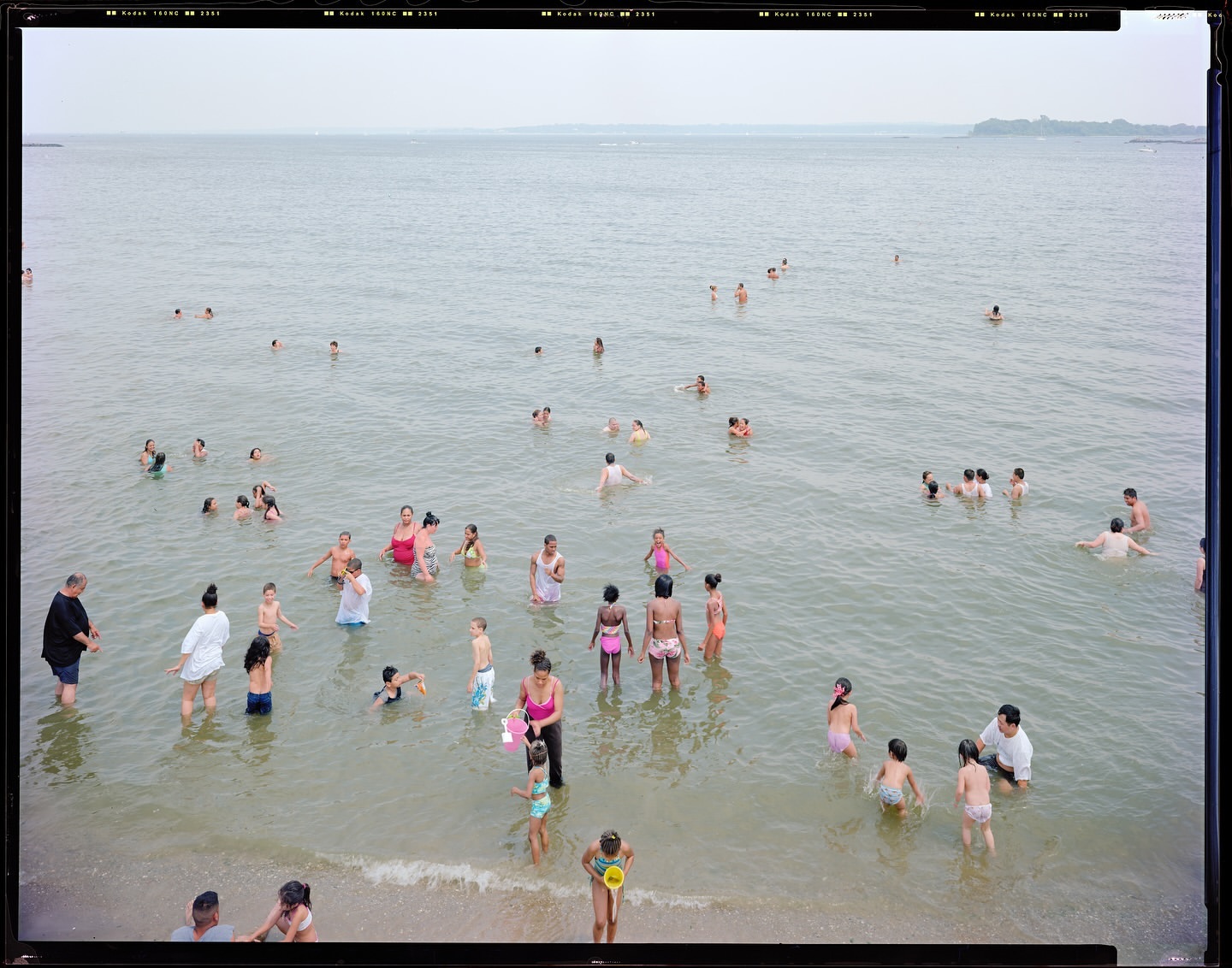 A crowded beach scene showing a large group of people wading and swimming in the ocean during the daytime on a bright sunny day.