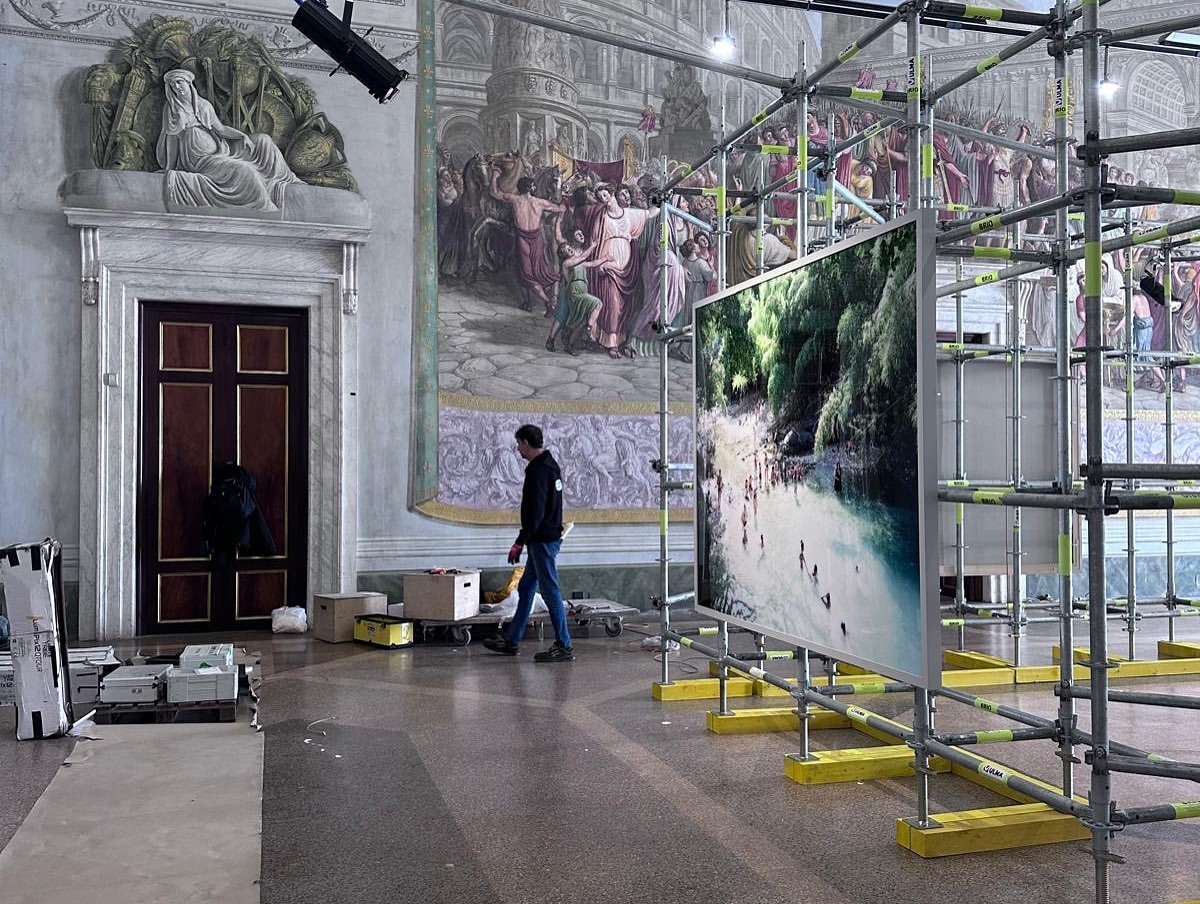 Man walks past a large framed photograph of a beach under construction scaffolding inside a historical building with painted walls.