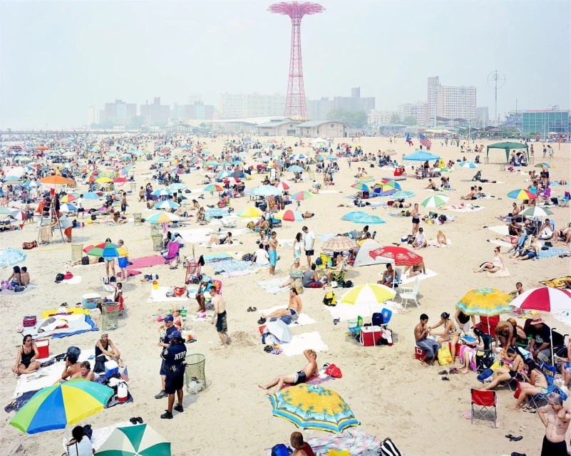 Aerial shot of a crowded Coney Island beach with numerous people, umbrellas, and the Parachute Jump in the distance on a hazy day.