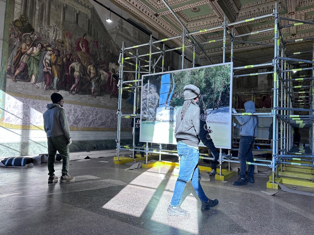 Indoor scene with three people installing photography art in a gallery space with high ceilings and a painted mural.