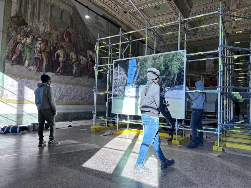 Indoor scene with three people installing photography art in a gallery space with high ceilings and a painted mural.