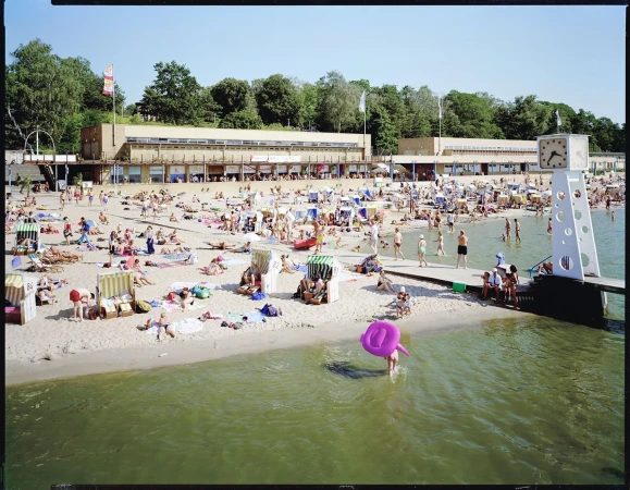Crowded beach scene with many people sunbathing, swimming, and relaxing near a large building and clock tower on a sunny day.