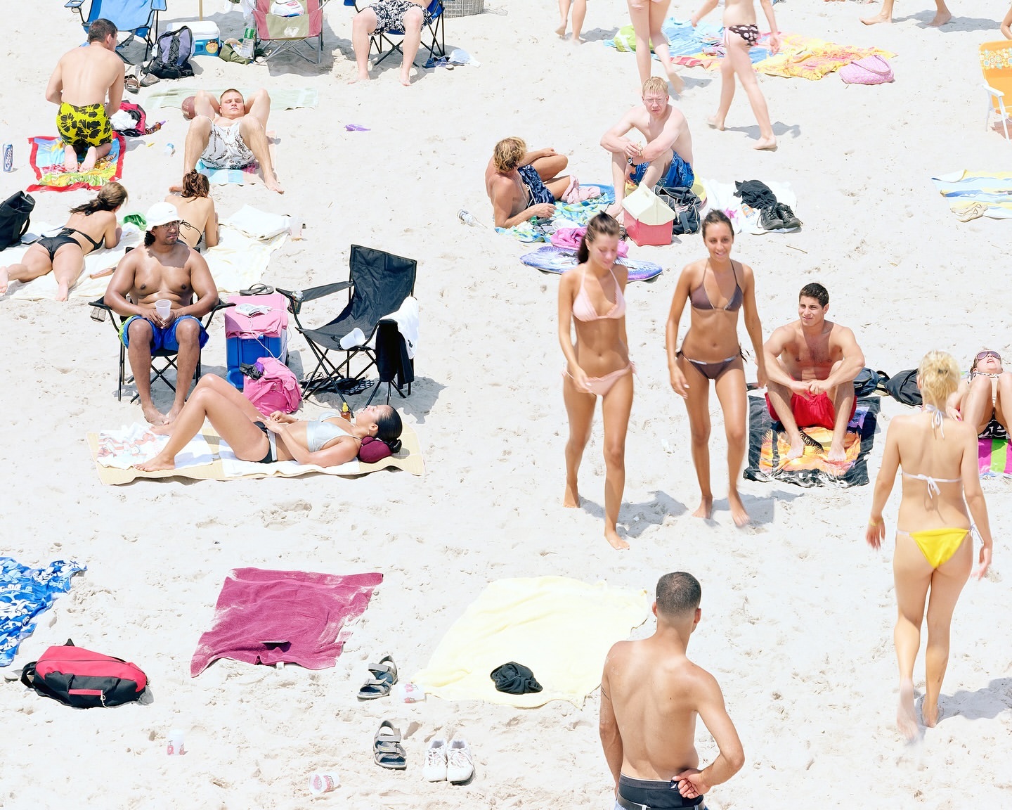 High-angle view of a crowded beach with many people in swimwear. Some are walking, some are sitting, some are laying down on towels.