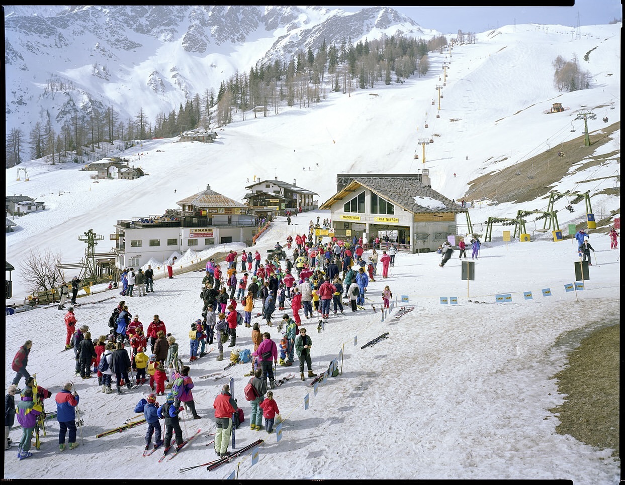 Large group of people skiing in a snow-covered mountain landscape with ski lifts and resort buildings in the background on a sunny day.