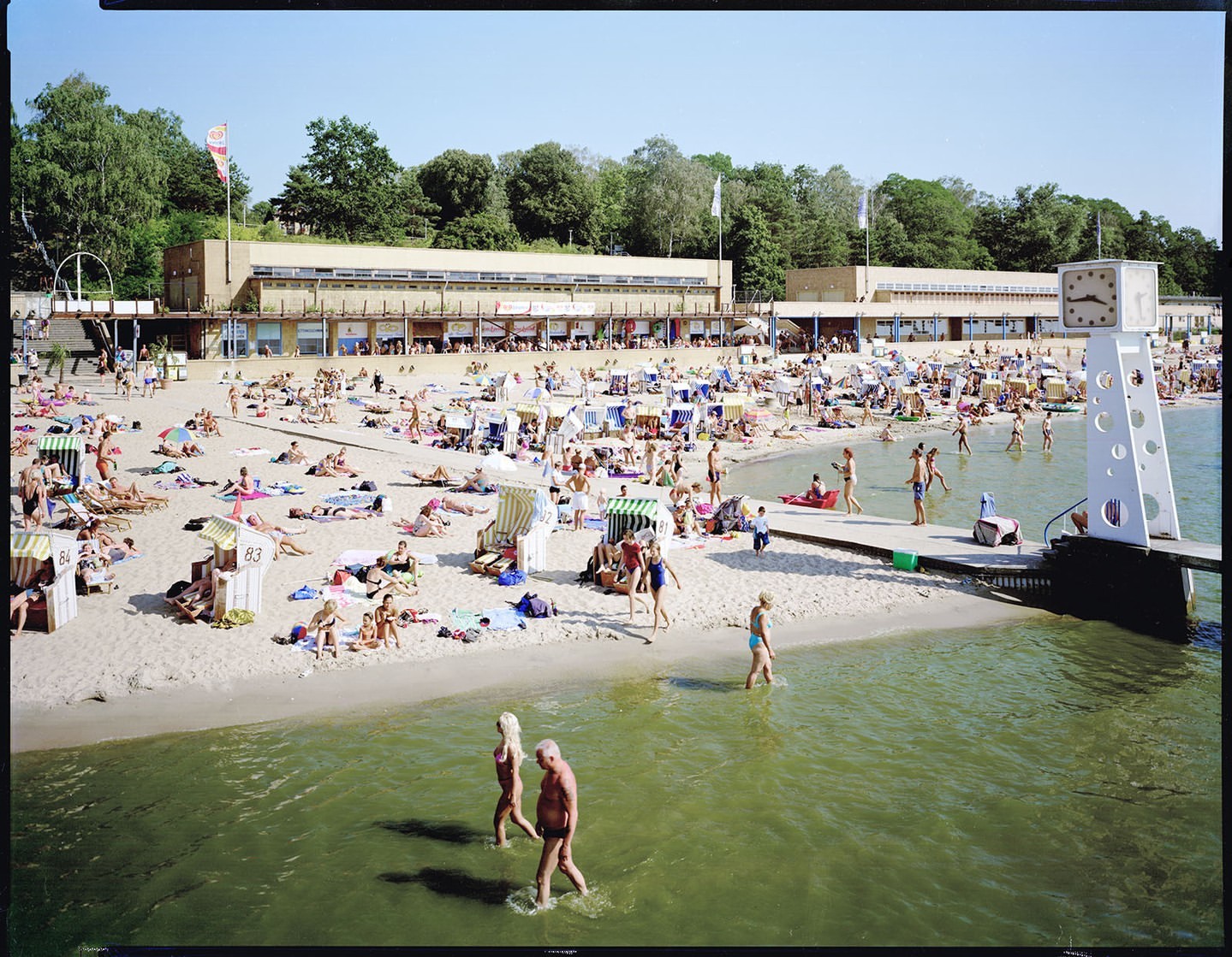 Sandy beach scene teeming with people sunbathing, swimming, and walking along the shore near modern building on a sunny day.
