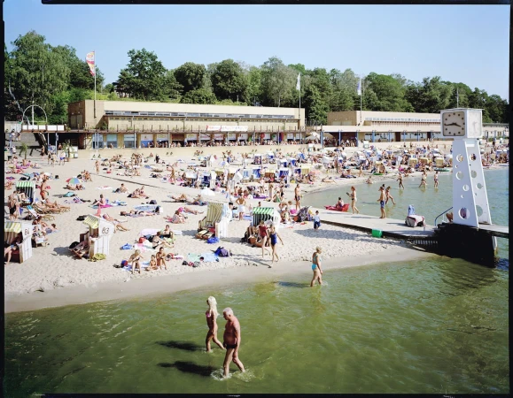 Sandy beach scene teeming with people sunbathing, swimming, and walking along the shore near modern building on a sunny day.