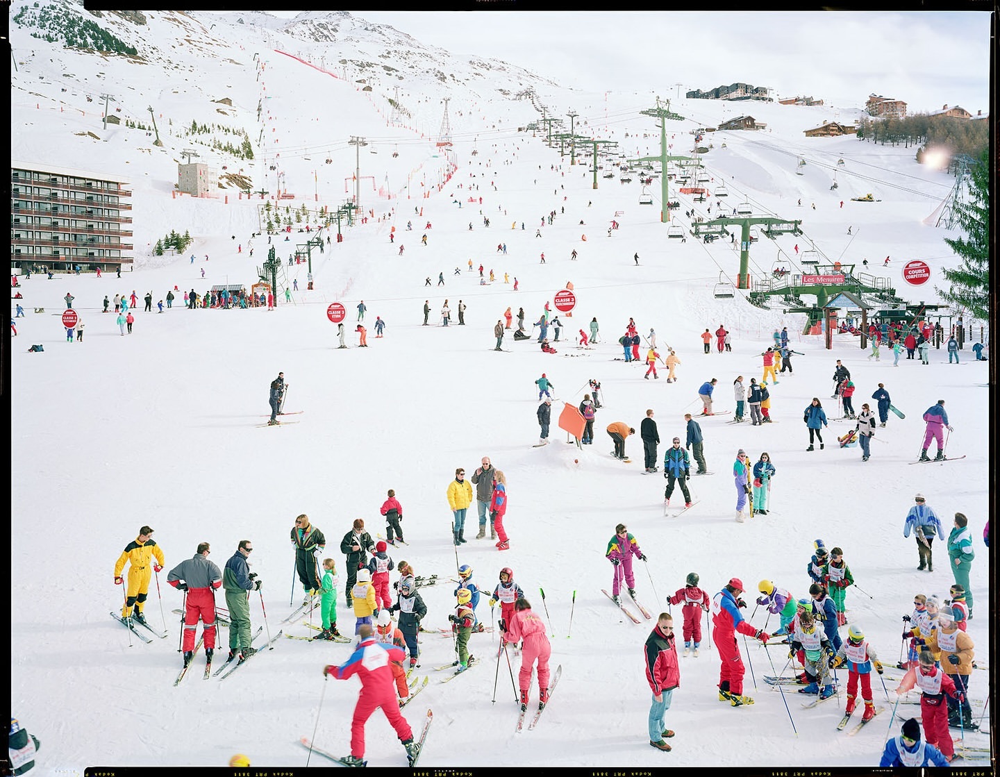 Large ski resort scene with groups of skiers and snowboarders on snow-covered slopes, mountain backdrop, ski lifts, and buildings.