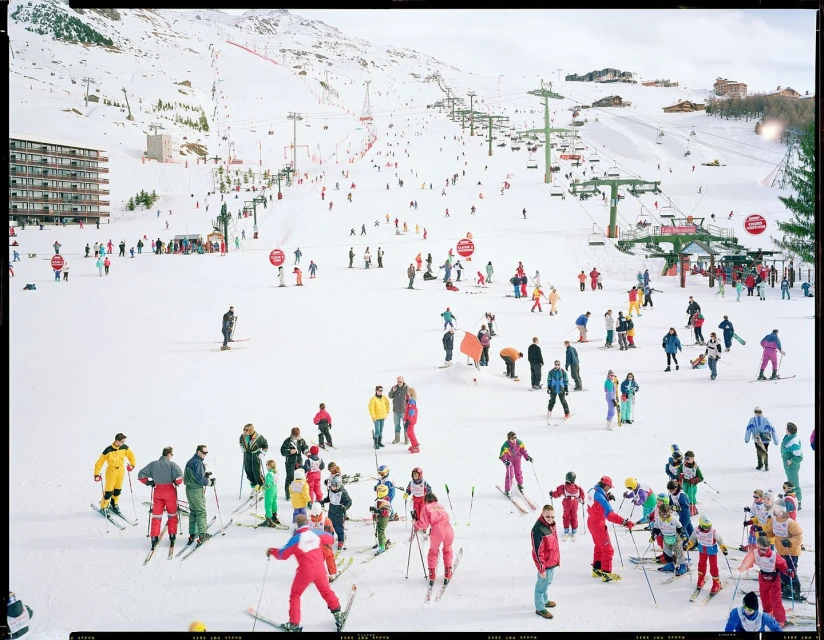 Large ski resort scene with groups of skiers and snowboarders on snow-covered slopes, mountain backdrop, ski lifts, and buildings.