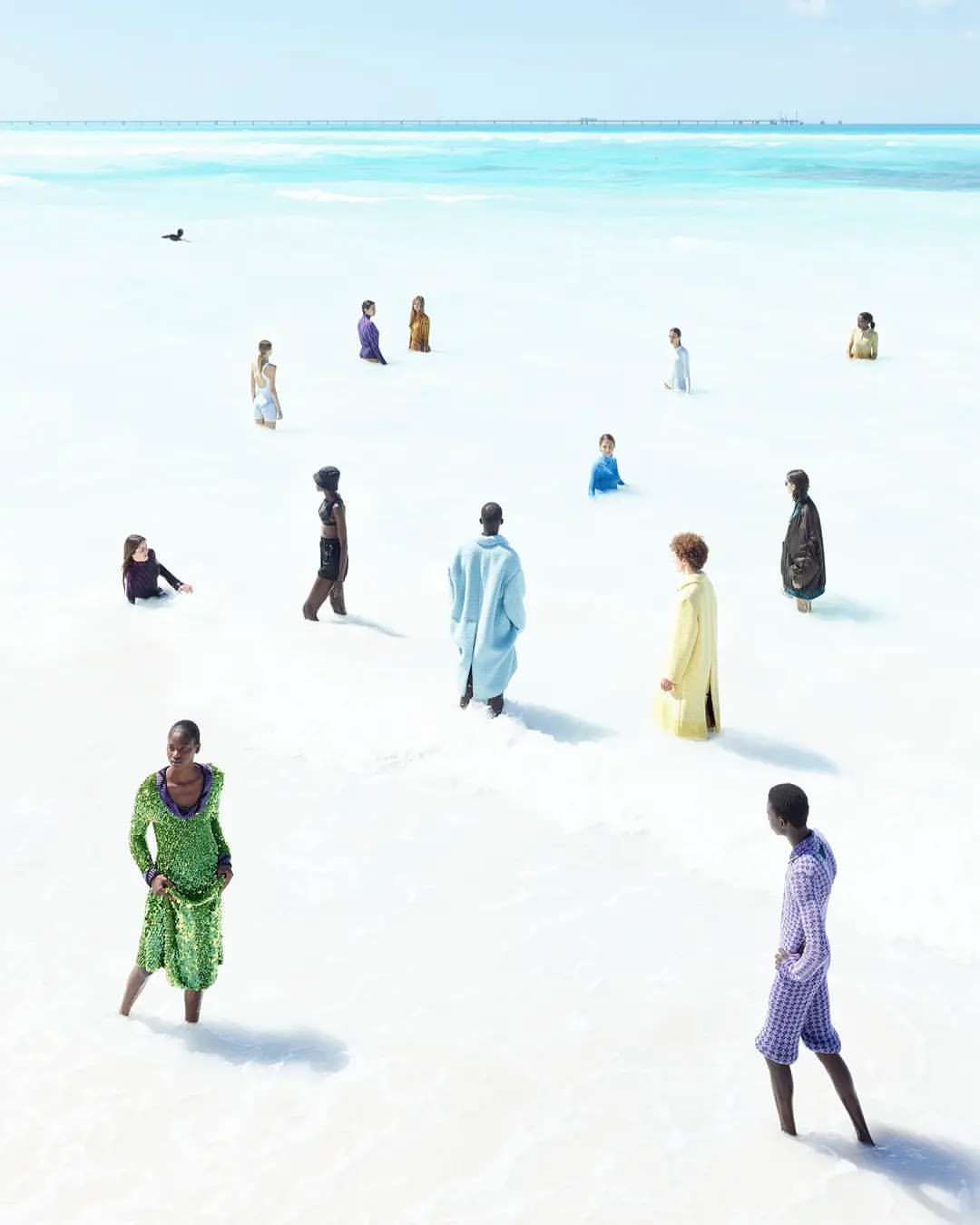 Fashion photography featuring a group of diverse models standing in shallow, clear water on a sunny day under a bright blue sky.