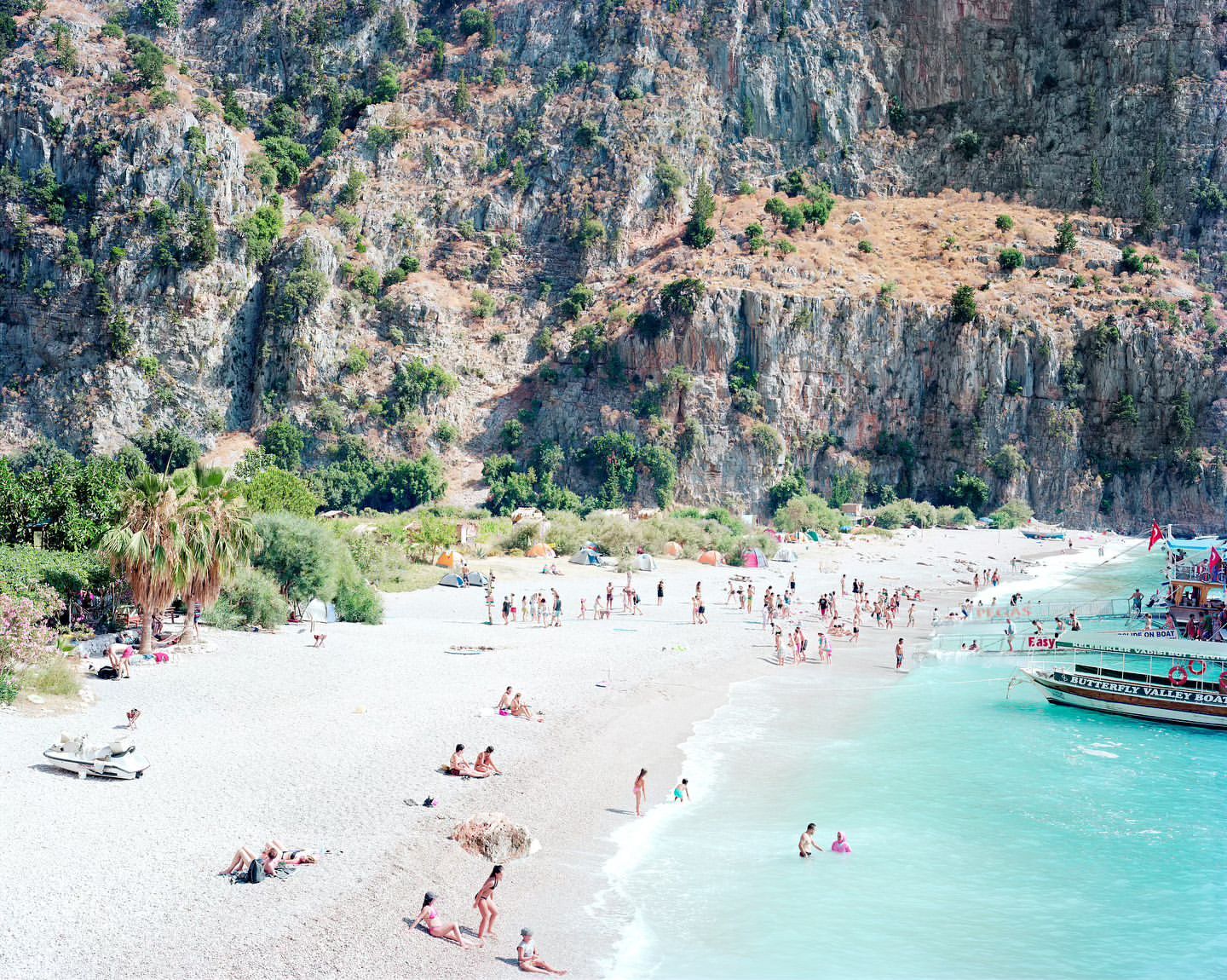 Aerial view of a beach with many people sunbathing and swimming near large cliffs and a boat in turquoise water on a sunny day.