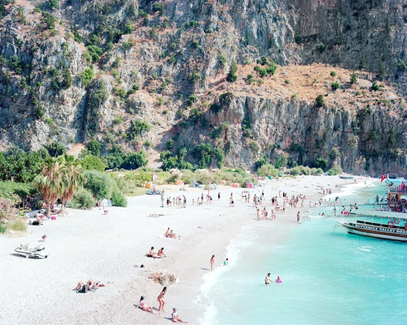 Aerial view of a beach with many people sunbathing and swimming near large cliffs and a boat in turquoise water on a sunny day.