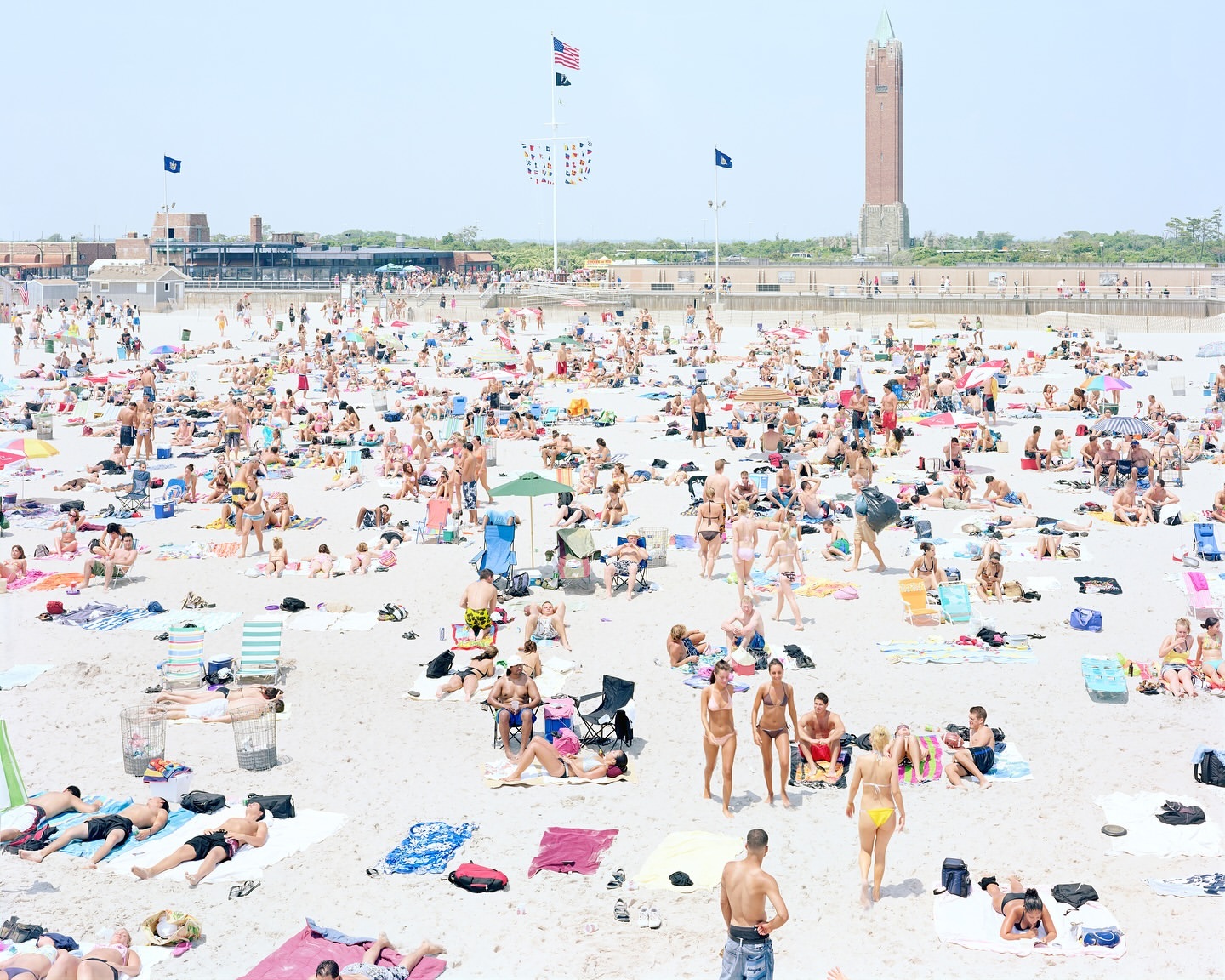High-angle view of a crowded sandy beach with many people sunbathing, swimming, and relaxing near a brick building and tower under a clear sky.