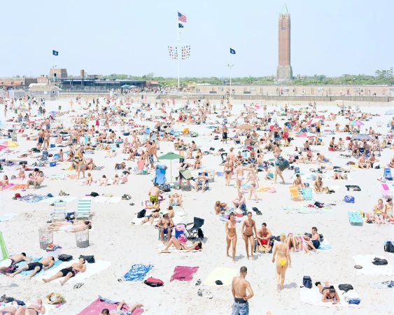 High-angle view of a crowded sandy beach with many people sunbathing, swimming, and relaxing near a brick building and tower under a clear sky.