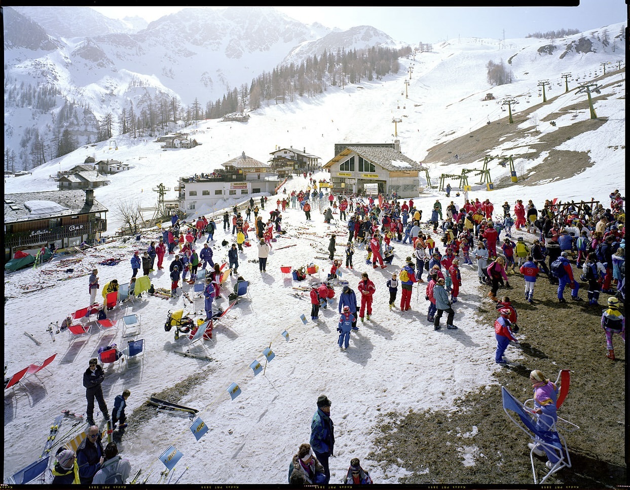 Large group of people gathering at a ski resort near buildings on a snowy mountain with chair lifts visible in the background.
