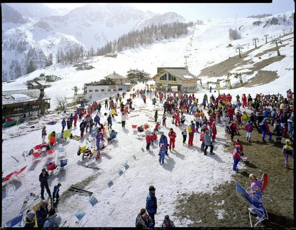 Large group of people gathering at a ski resort near buildings on a snowy mountain with chair lifts visible in the background.