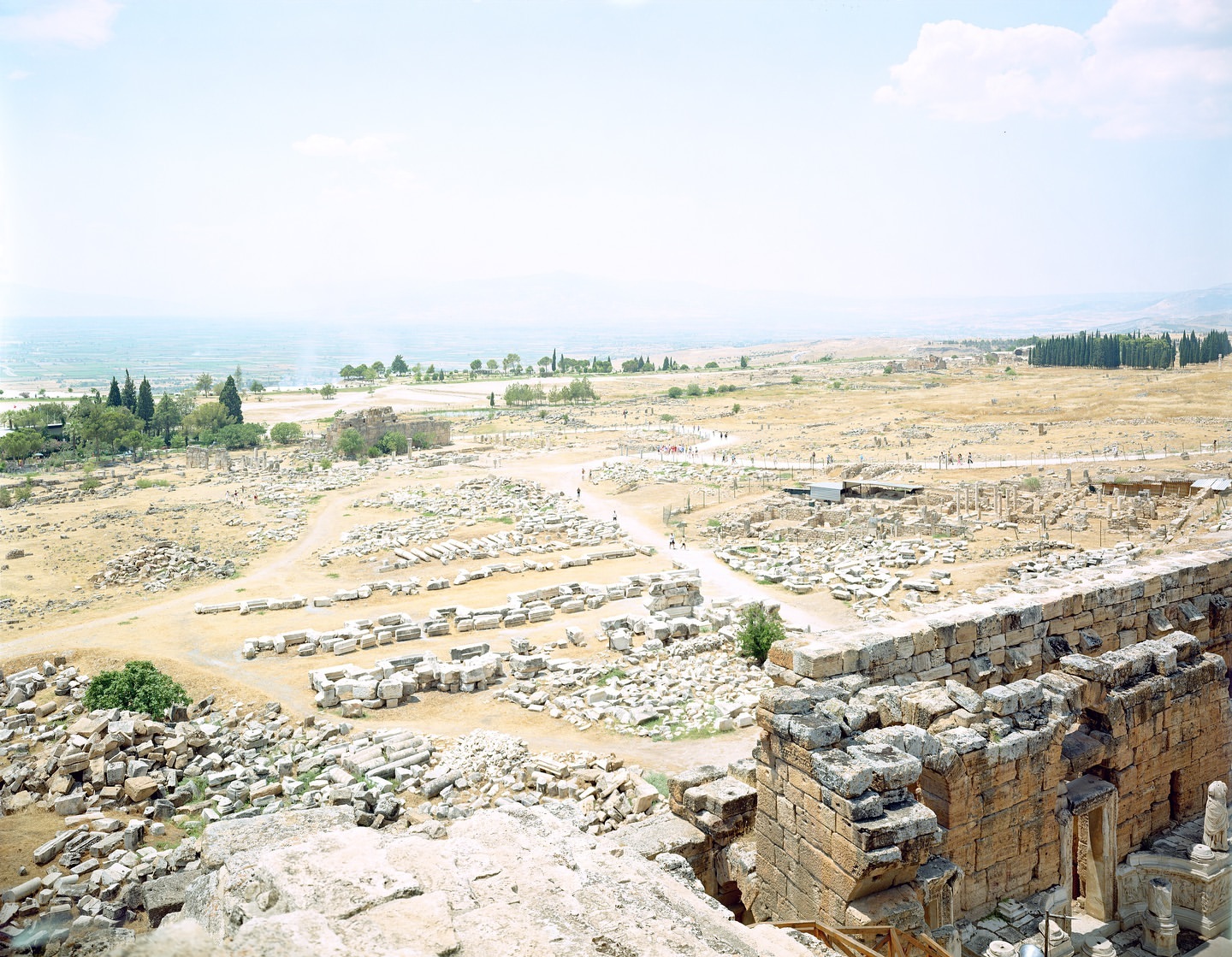 Aerial view of ancient stone ruins in a desert landscape, featuring an old stone wall, trees, a distant ocean, and a clear sky.