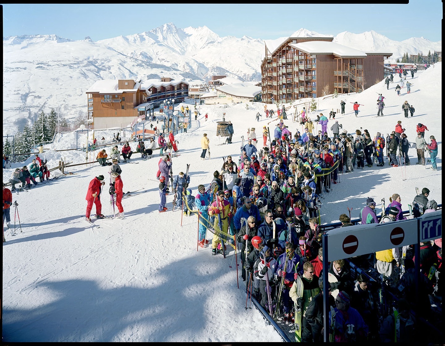 Large group of skiers waits in line at a mountain resort, with snow-covered slopes and buildings in the scenic background on a sunny day.