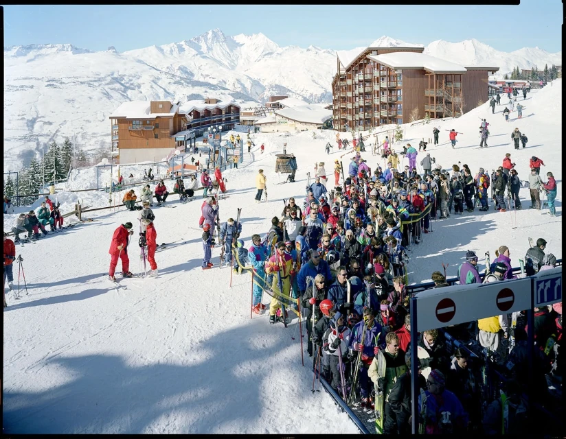 Large group of skiers waits in line at a mountain resort, with snow-covered slopes and buildings in the scenic background on a sunny day.