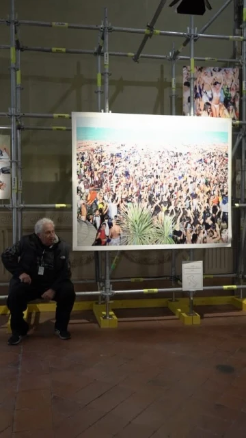An older man sits on a bench in front of a large photograph of a crowded beach scene at an art gallery exhibit.
