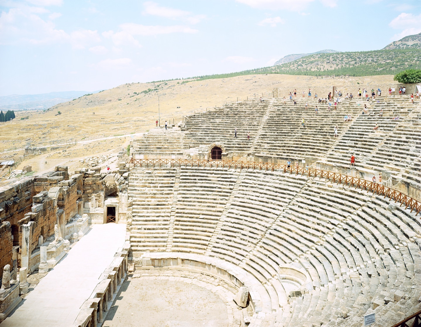 Vast ancient amphitheater with stone seating and partial structure, surrounded by dry grassy hills under a bright, sunny sky.