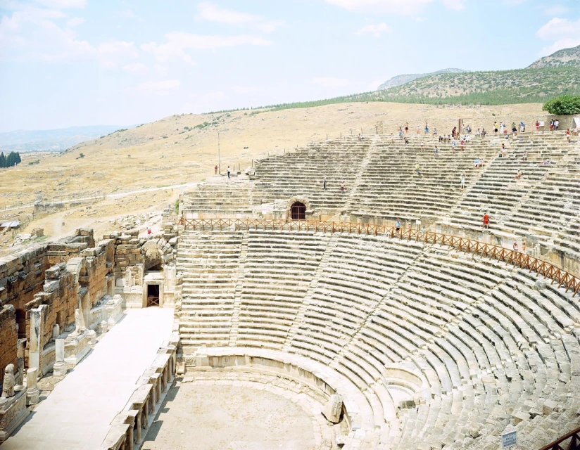 Vast ancient amphitheater with stone seating and partial structure, surrounded by dry grassy hills under a bright, sunny sky.