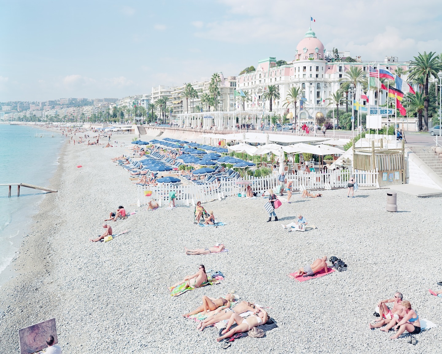 Overhead shot of sunbathers on a pebble beach in Nice, France, with turquoise water, colorful umbrellas, and a grand hotel in the background.