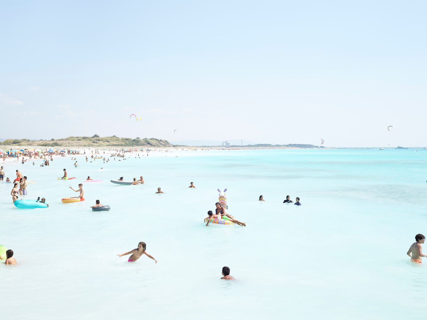 Crowd of people enjoying a sunny day at the beach, swimming and wading in the turquoise water near the white sand.