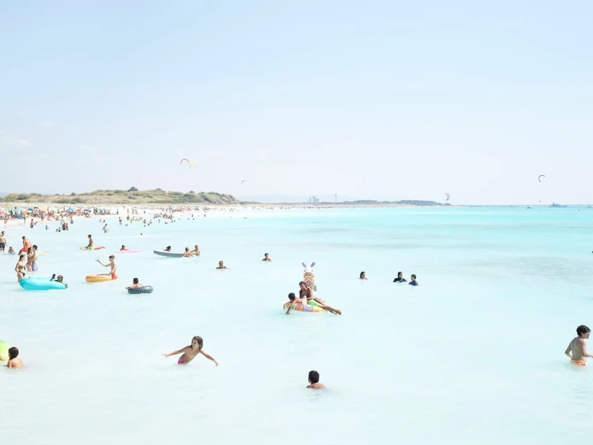 Crowd of people enjoying a sunny day at the beach, swimming and wading in the turquoise water near the white sand.