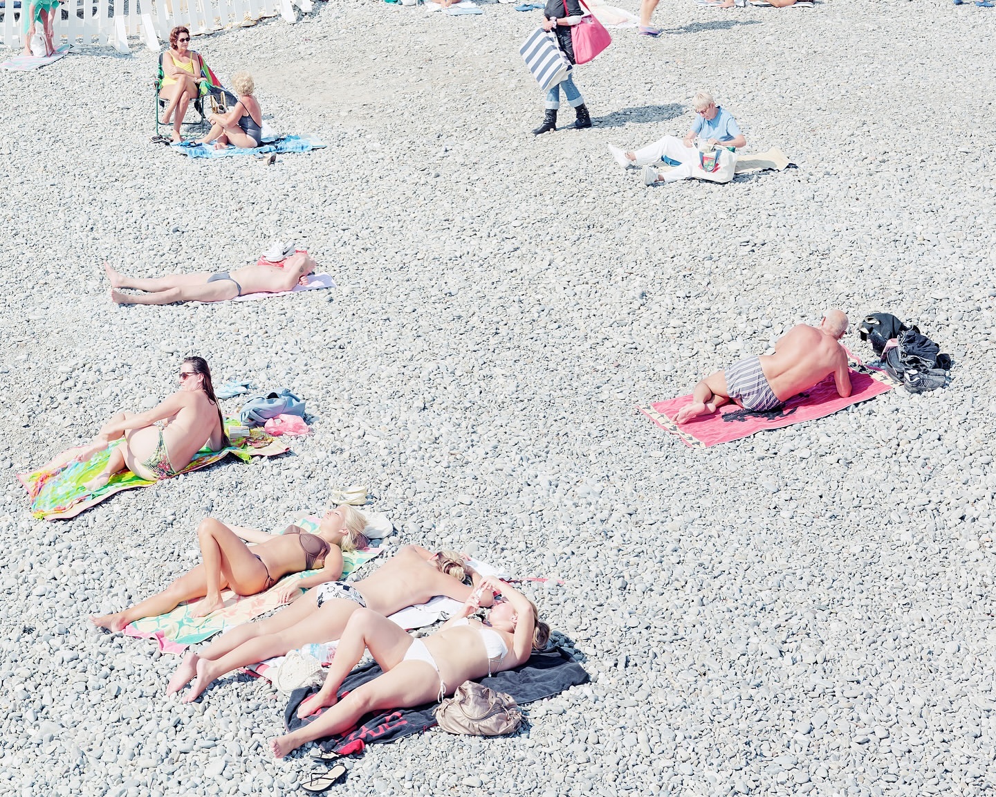 High-angle view of people sunbathing and relaxing on a pebble beach, enjoying the summer heat near the water's edge on a sunny day.