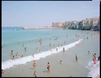 High-angle view of a crowded beach and shallow turquoise ocean with numerous people swimming and wading under a clear sky.