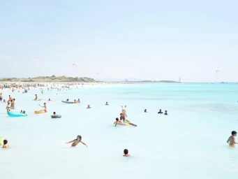 Wide panoramic beach scene with numerous people swimming and relaxing in turquoise water under a pale blue sky.